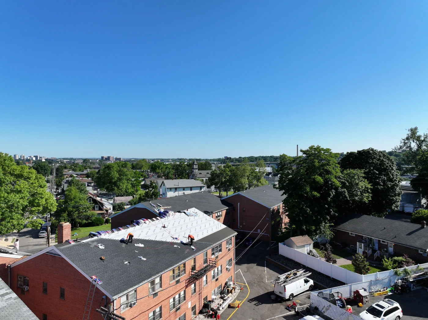 A professional crew installs white underlayment and asphalt shingles on a large multi-family brick building complex, demonstrating a comprehensive commercial roofing project under a clear blue sky.