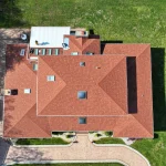 Aerial view of a red shingle commercial roof with skylights, crews working near a flat roof area, lawn, and paved driveway.