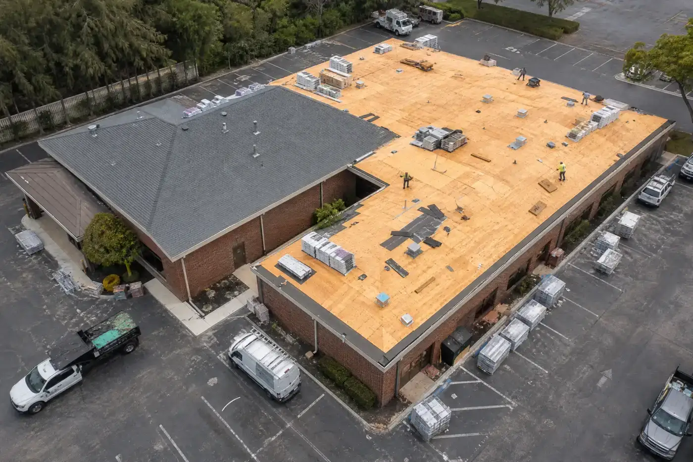 An L-shaped business in the process of getting a roof replacement. Part of the roof is covered in gray shingles, and other parts have the wooden deck exposed. Roofing materials can be seen on the rooftop and staged in the parking lot, which surrounds the building on three sides.