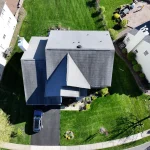 Aerial view of a gray shingle roof on a suburban home with driveway, lawn, neighboring houses, and roofing crews nearby.