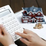 A close-up of a person’s hands holding and using a white calculator. In the background of the image, a dark wooden table has a clipboard with multiple papers resting atop it. On top of the clipboard, a pen, some coins, and a model of a red home with a dark gray roof can be seen.