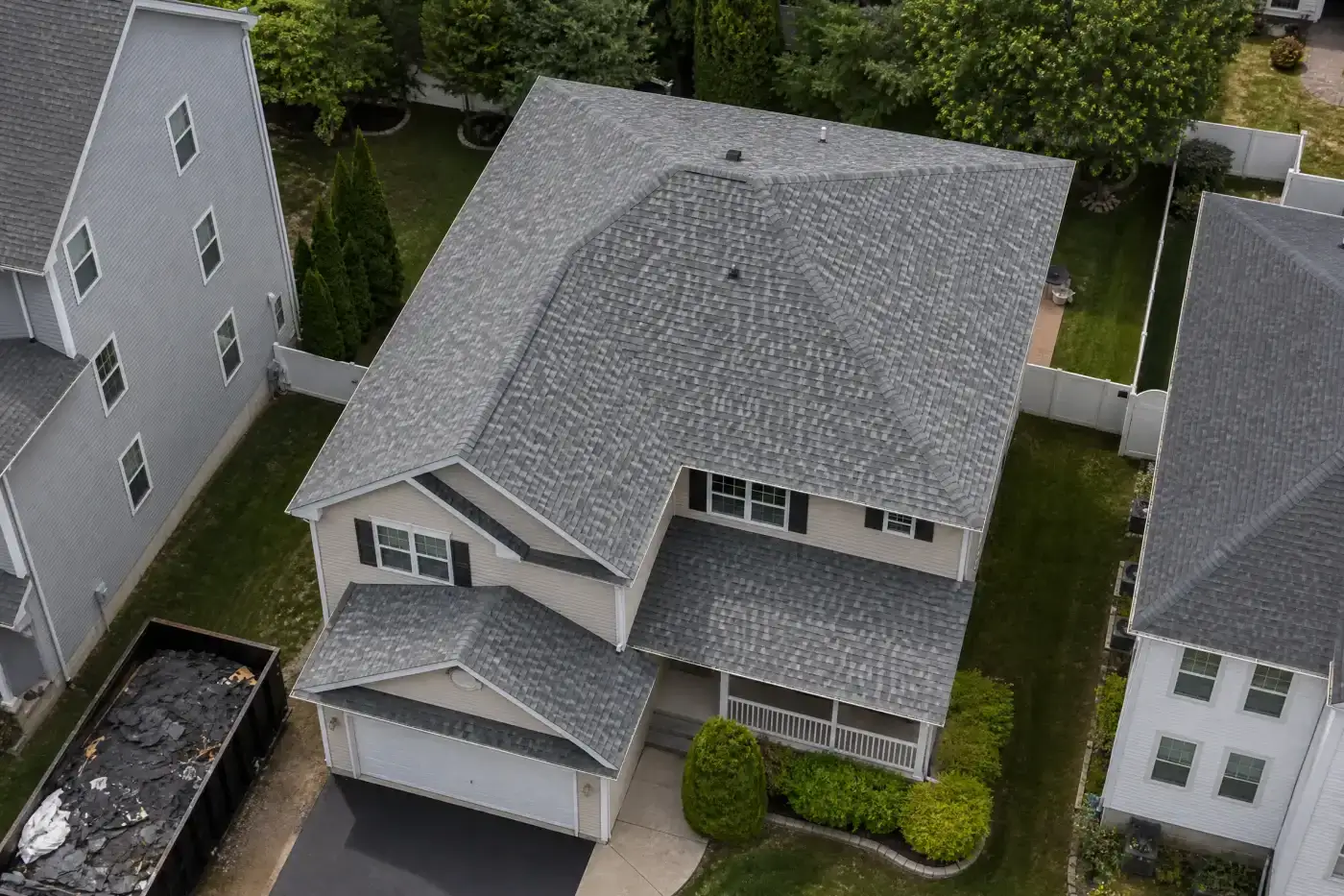 A photo taken of a multi-level home’s roof using a drone. The roof is between two other, taller homes, and has shingles in varied shades of gray covering the surface. At the bottom left of the image, a dumpster filled with roofing materials can be seen.