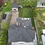 Aerial view of a completed gray shingle roof on a large home with chimneys, skylight, driveway, and backyard.