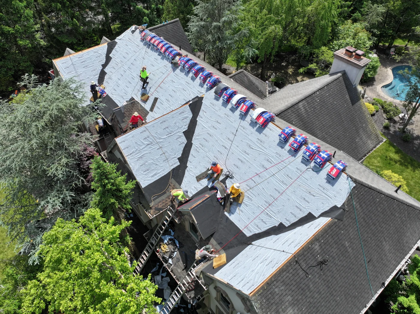 An aerial view captures a professional installation crew carefully laying down protective underlayment and staging bundles of architectural shingles across a multi-gabled residential roof, showcasing a major project managed by an expert roofing contractor.