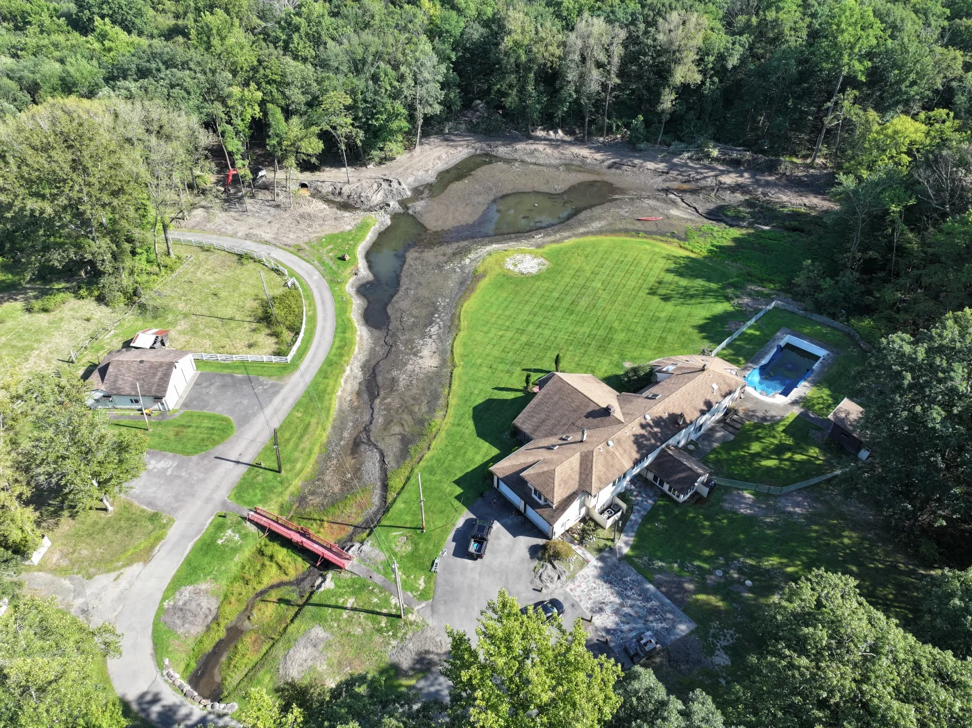 A wide aerial view captures a sprawling residential property featuring a large home with a brown shingle roof, a swimming pool, and an expansive lawn next to a winding stream bed, showcasing a pristine installation by a superior roofing contractor.
