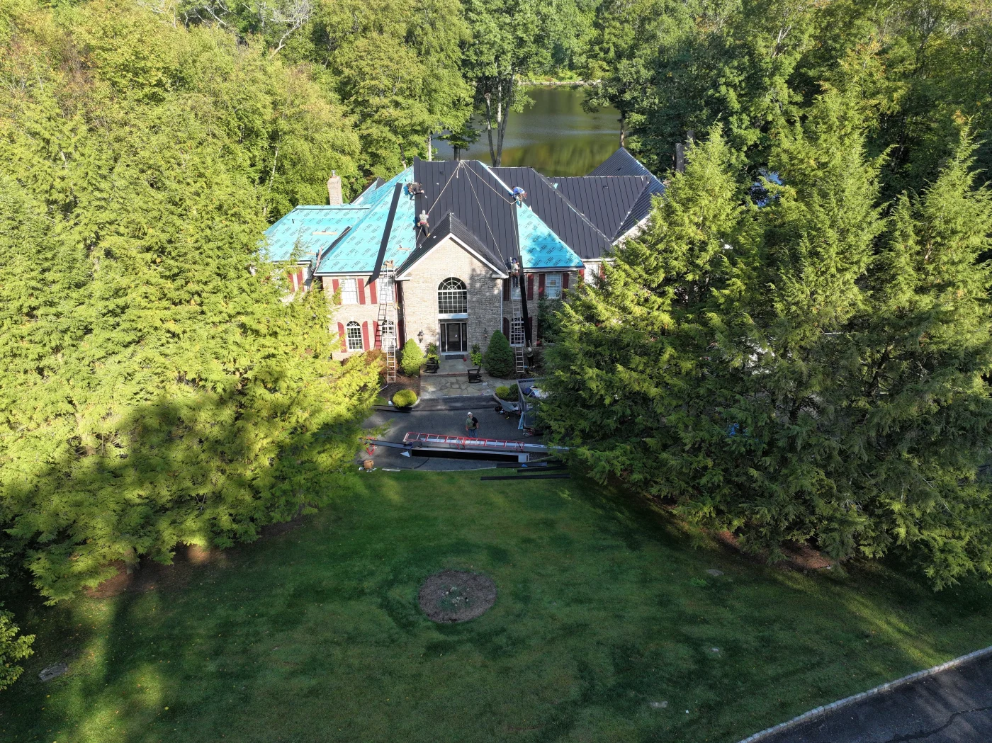 Top-Notch Roofing Contractor | Boonton, NJ | Reisch Roofing & Construction An aerial drone photograph captures a large brick estate surrounded by dense trees as a professional crew installs a modern black metal roof over blue underlayment, showcasing the precise work of a top-notch roofing contractor.