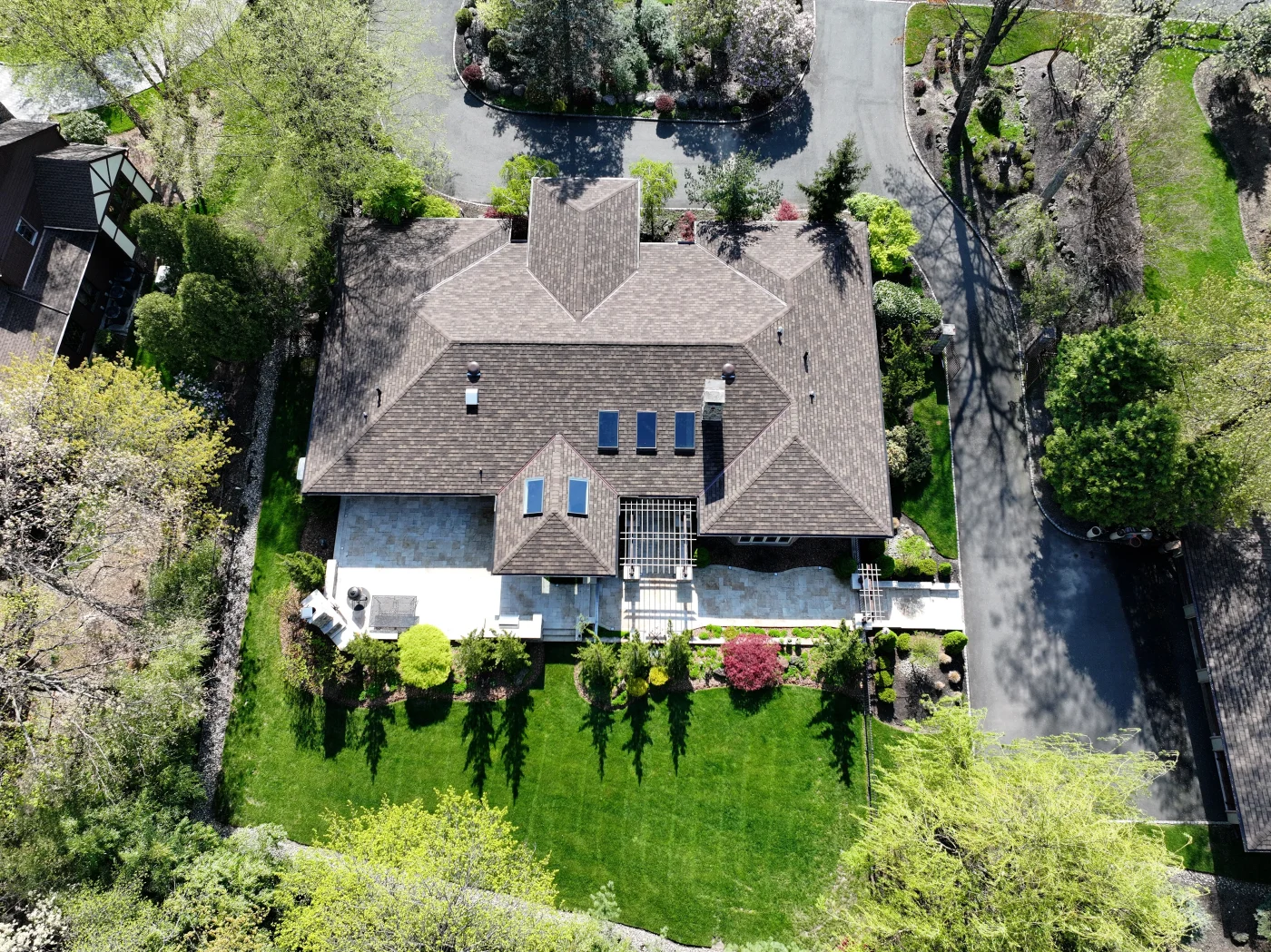 An aerial top-down view captures a luxurious estate with a sprawling brown architectural shingle roof, skylights, and a beautifully landscaped stone patio and lawn that showcase the quality of professional roofing services.